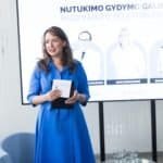 Ryškiai mėlyna suknele vilkinti moteris kalba laikydama planšetinį kompiuterį ir stovėdama priešais ekraną, kuriame rodoma informacija apie renginį ir trijų žmonių nuotraukos. Aplinka atrodo moderni ir profesionali, šalia stovi kėdės ir stiklinės vandens.