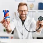 Baltu chalatu vilkintis gydytojas vienoje rankoje laiko žmogaus širdies modelį, kitoje - stetoskopą, o biuro fonas neryškus.