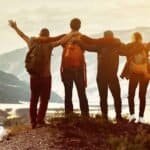 Keturi žmonės su kuprinėmis stovi ant kalno viršūnės, susikibę rankomis ir iškėlę rankas į viršų, žvelgdami į vaizdingą kalnų kraštovaizdį saulėlydžio metu.