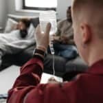 Raudonais drabužiais vilkintis medicinos darbuotojas laiko lašelinę, ruošdamas ją lovoje gulinčiam pacientui, o kitas asmuo sėdi šalia, kambaryje su švelnia natūralia šviesa.