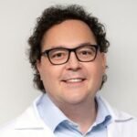 Besišypsantis žmogus garbanotais rudais plaukais, dėvintis juodus akinius, šviesiai mėlynus marškinius su apykakle ir baltą laboratorinį chalatą, stovintis vienspalviame šviesiame fone.