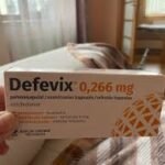 Jaukioje miegamojo aplinkoje su lova ir langu fone ranka laiko "Defevix" 0,266 mg kapsulių dėžutę.