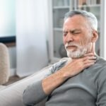 Vyresnio amžiaus vyras žilais plaukais ir barzda sėdi ant sofos, užmerktomis akimis ir nepatogiai nusiteikęs, laikydamas ranką prie gerklės, o tai rodo, kad jam skauda gerklę arba sunku ryti.