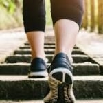 Stambiu planu - asmuo, avintis juodus sportinius batelius ir mūvintis juodas kelnes, einantis lauko akmeniniais laiptais, šonuose - žaluma, o fone - saulės šviesa.