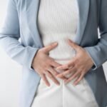 Šviesiai mėlyną švarką ir baltus marškinius vilkintis asmuo abiem rankomis laiko pilvą ir atrodo, kad jaučia pilvo diskomfortą.