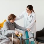 Sveikatos priežiūros specialistas padeda pagyvenusiai moteriai, naudojančiai vaikštynę, atsistoti nuo lovos. Pagyvenusi moteris dėvi akinius ir pilką megztinį; sveikatos priežiūros specialistas vilki baltą chalatą su stetoskopu.