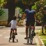 Suaugęs ir vaikas važiuoja dviračiais vienas šalia kito saulėtu, medžiais apsodintu dviračių taku, apsuptu žydinčių krūmų ir žalumos. Nuotrauka daryta iš nugaros, kai jie važiuoja tolyn nuo fotoaparato.