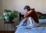 Moteris sėdi ant lovos, pritraukusi kelius prie krūtinės, ir atrodo susimąsčiusi arba liūdna. Ji vilki rudą megztinį ir baltą sijoną, o šalia jos ant lovos stovi vazoninis augalas ir telefonas.