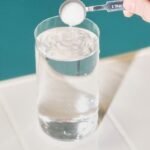 Ranka laiko šaukštą baltų miltelių virš skaidraus stiklinės vandens, tarsi norėdama miltelius įpilti į vandenį. Fonas yra žalsvai mėlynas, o paviršius – baltas.