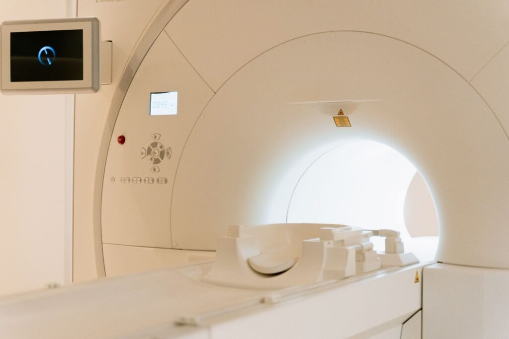Medicinos įstaigoje esančio MRT aparato artimas vaizdas, kuriame matoma apvali anga ir valdymo pultas su mygtukais bei ekranu kairėje sienoje.