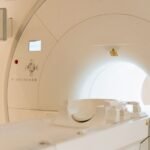 Medicinos įstaigoje esančio MRT aparato artimas vaizdas, kuriame matoma apvali anga ir valdymo pultas su mygtukais bei ekranu kairėje sienoje.