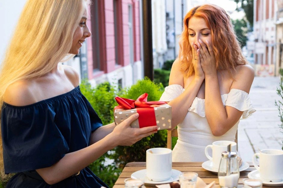 Dvi moterys sėdi lauko kavinės staliuke; viena iš jų paduoda kitai dovaną su raudonu kaspinu, o ši atrodo nustebusi ir laiminga; priešais jas stovi kavos puodeliai ir pyragaičiai.